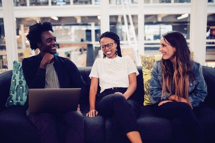 One male Black student, a female Black student, and a white female smiling and laughing while sitting on a grey couch in the lobby of a university.