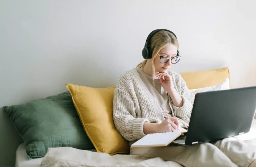 blond girl with headphones at laptop taking notes
