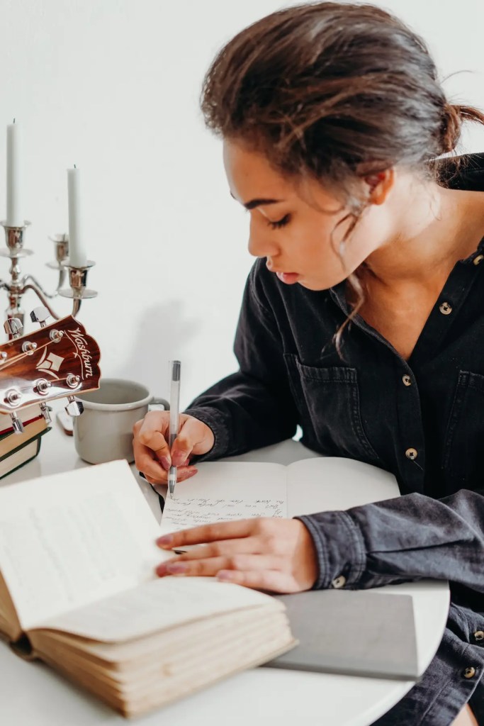 Focused student writing in a notebook at a desk with books and a guitar nearby—capturing the personal and reflective process of crafting a Master of Public Policy statement of purpose.