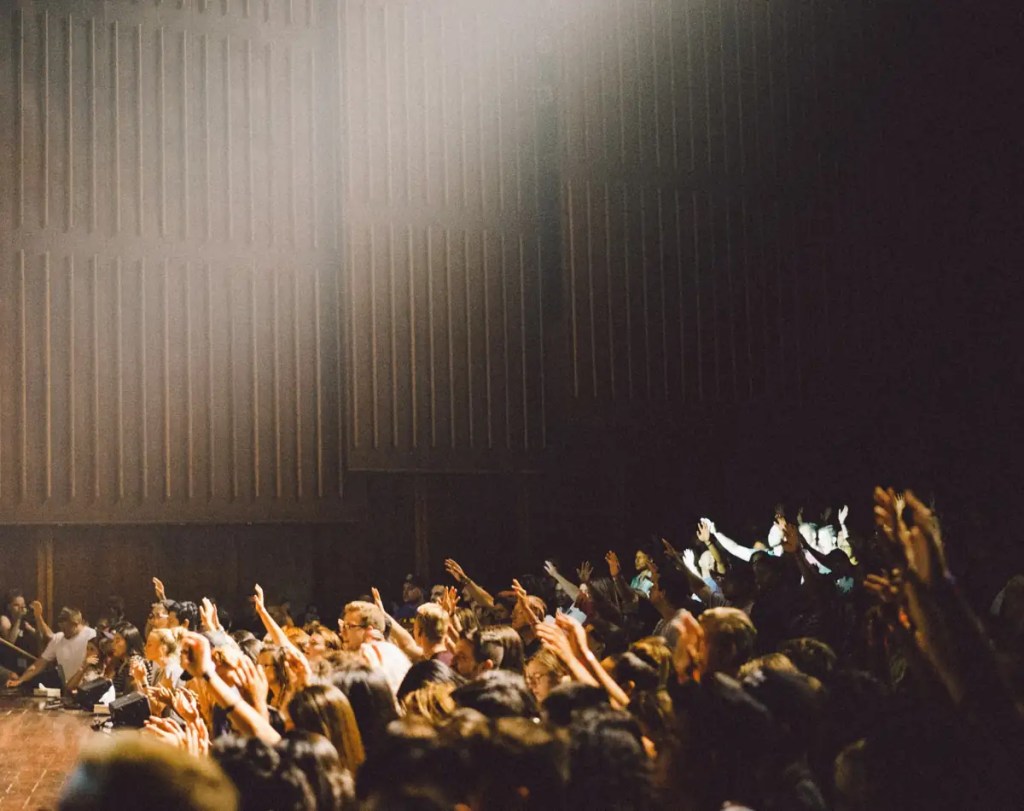 A large group of people raising their hands in a dark auditorium under a spotlight.