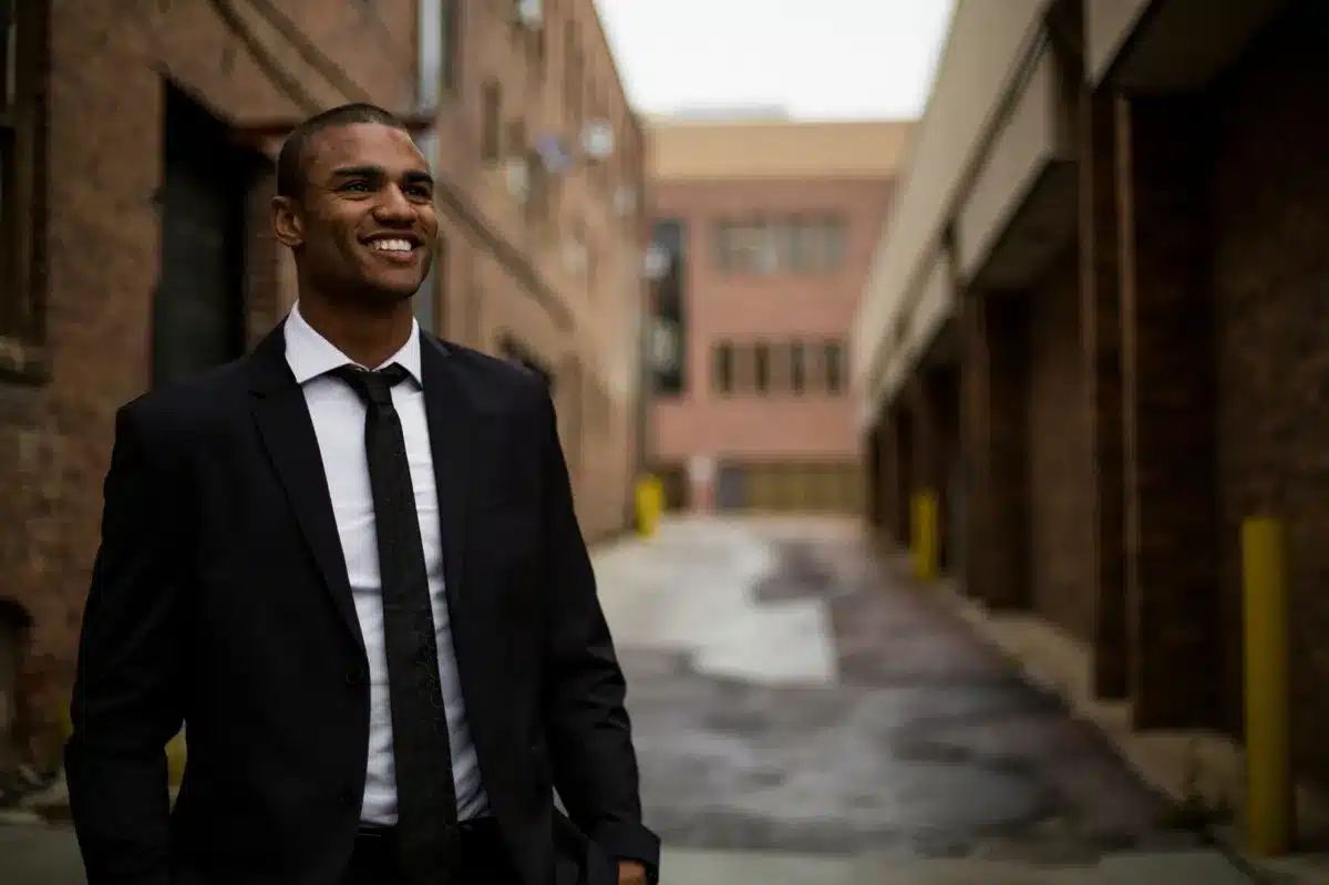 Smiling man in a suit standing confidently in an urban alleyway between brick buildings.
