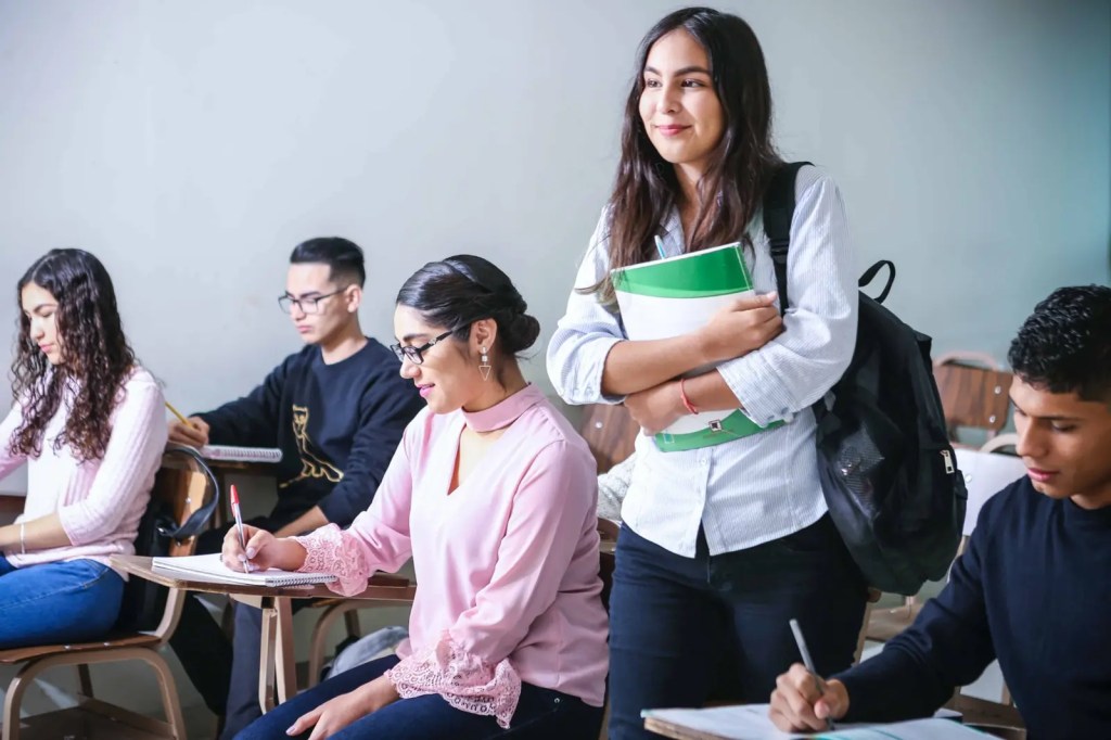 Group of diverse students sitting in a classroom, with one female student standing and smiling while holding a folder.