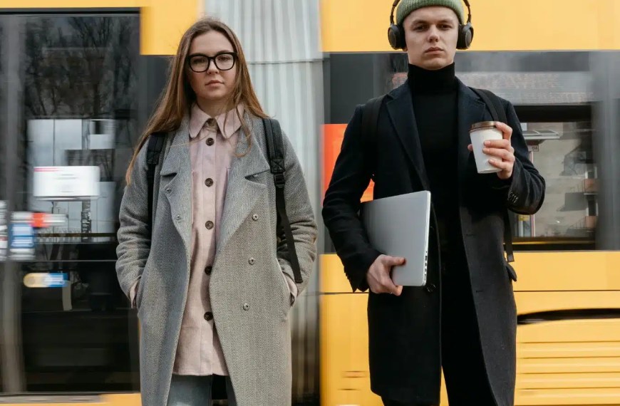Two young professionals stand on a train platform, one holding a laptop and coffee, the other with hands in pockets, as a yellow and red train passes behind them.