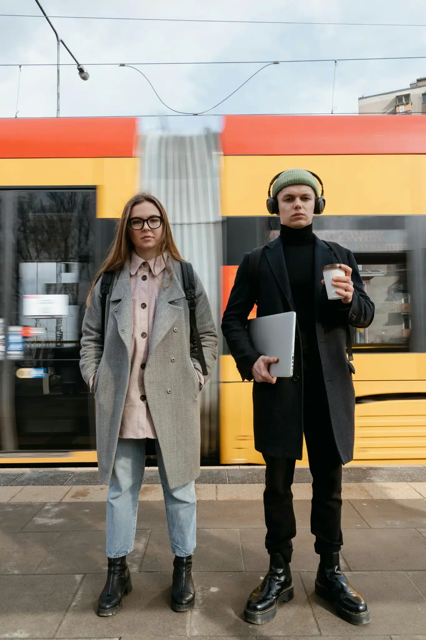 Two young professionals stand on a train platform, one holding a laptop and coffee, the other with hands in pockets, as a yellow and red train passes behind them.