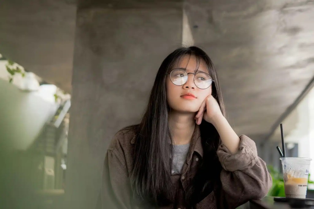 Young PhD student with long hair and glasses looking thoughtful while sitting at a table with an iced coffee.