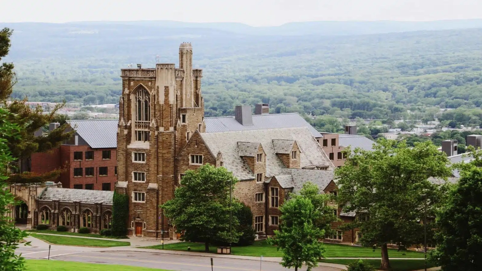 Gothic-style university building surrounded by trees with a forest-covered mountain range in the background.