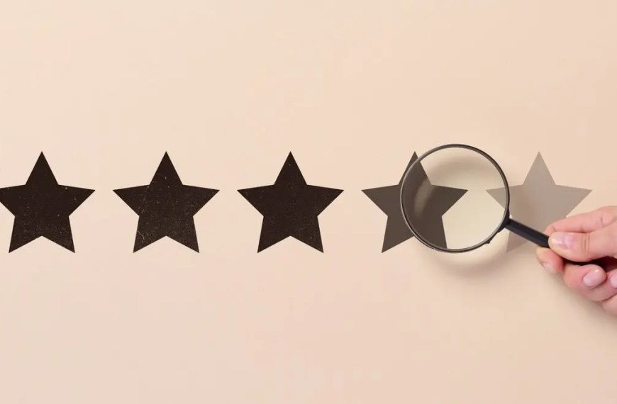 A hand holding a magnifying glass over black star ratings on a beige background, symbolizing careful evaluation of PhD program rankings and prestige.