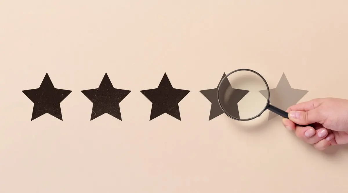 A hand holding a magnifying glass over black star ratings on a beige background, symbolizing careful evaluation of PhD program rankings and prestige.