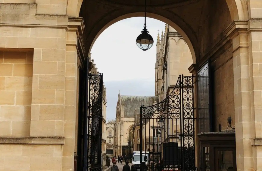 Stone archway entrance to a historic university campus with wrought iron gates and people walking inside