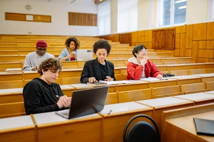 A diverse group of students seated in a lecture hall, some using laptops and taking notes, representing the GPA vs GRE decision-making process for graduate school admissions.