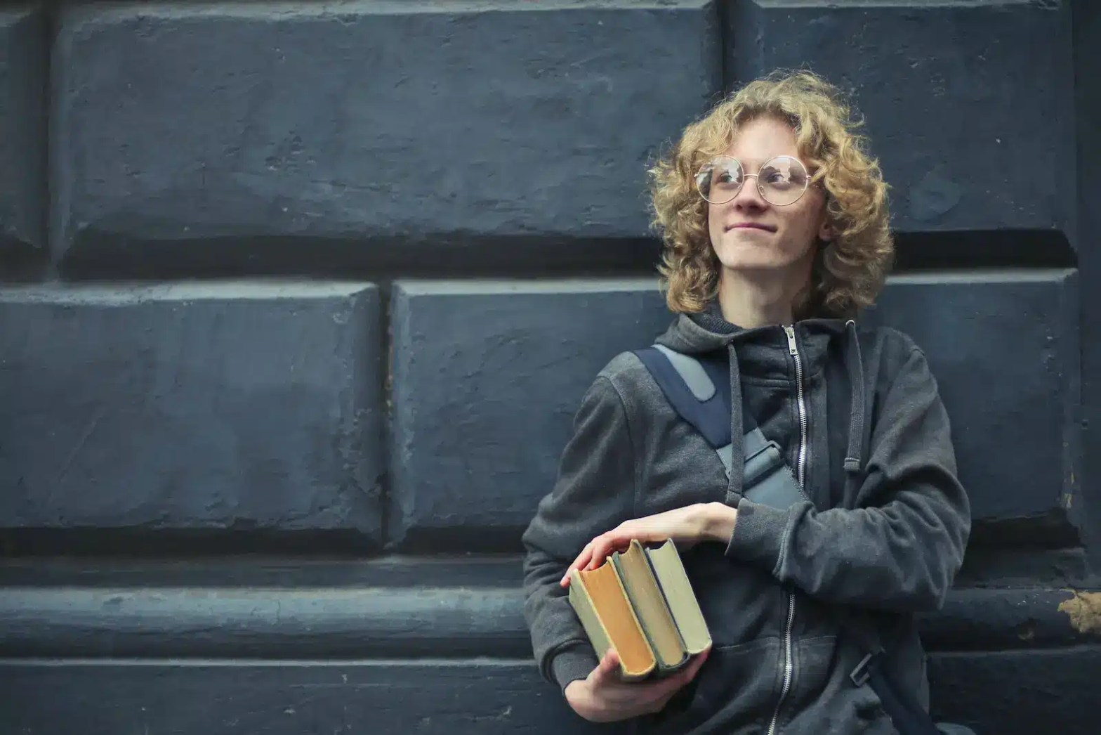 A student with curly hair and glasses wearing a dark hoodie and backpack, holding three books, standing against a stone wall, symbolizing the GPA vs GRE decision process for graduate school.