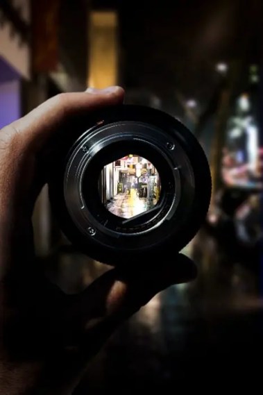 A hand holds a camera lens through which a sharp, well-lit alleyway is visible, symbolizing clarity and focus—ideal for a blog about how to write effectively about your research interests.